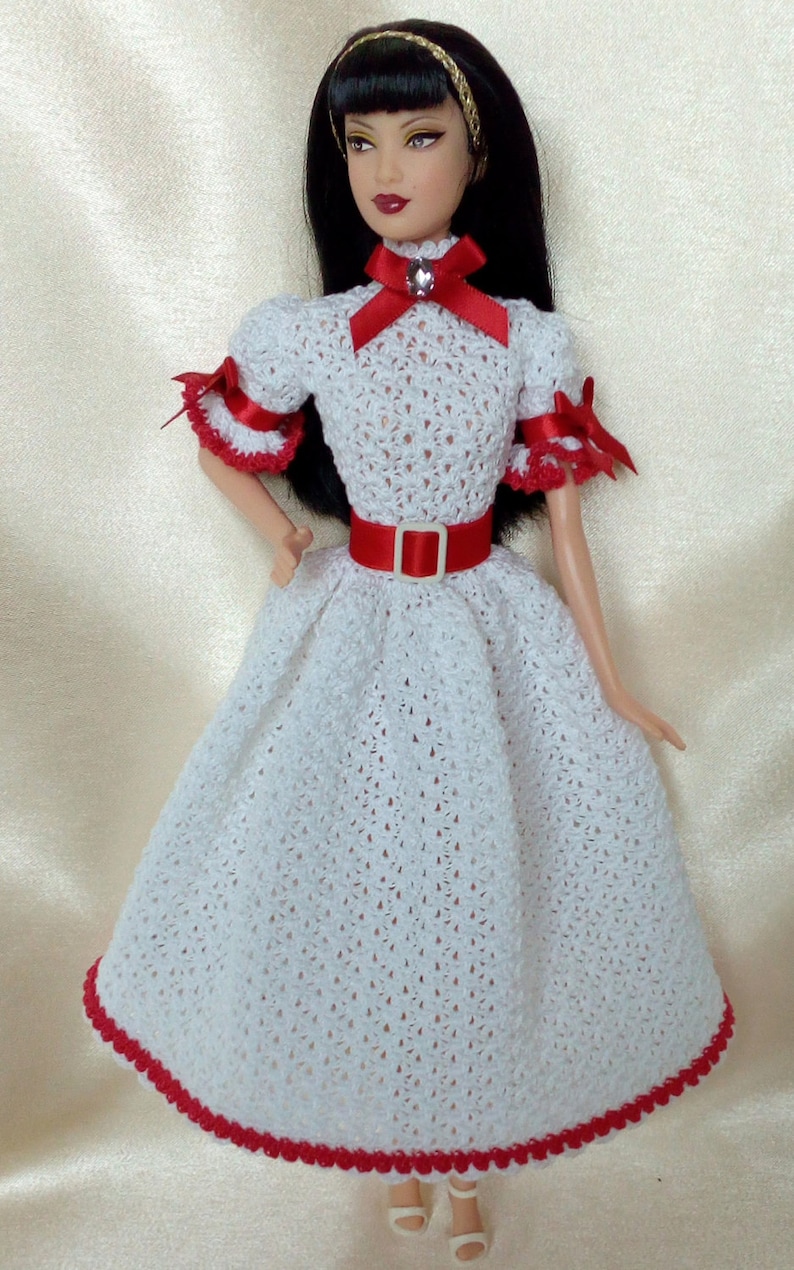 May include: A doll wearing a white crocheted dress with red trim and accents. The dress features a red belt and bow, and the doll has black hair and a gold headband. The dress is a vintage style.
