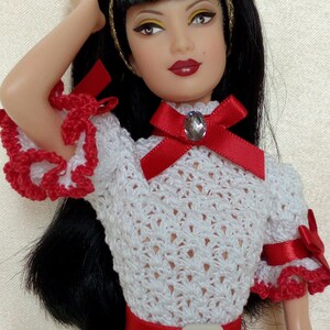 May include: A doll with black hair and a white crocheted dress with red accents. The dress features a red belt and bow with a gem. The doll has a gold headband and red lipstick.