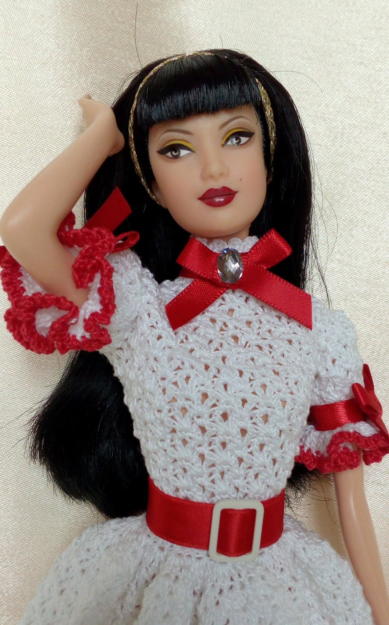 May include: A doll with black hair and a white crocheted dress with red accents. The dress features a red belt and bow with a gem. The doll has a gold headband and red lipstick.