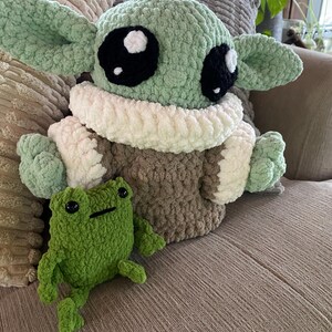 May include: A large, crocheted plush figure of a character with large black eyes, green skin, and large ears, wearing a brown and white outfit. A small green crocheted frog sits in front of the figure.