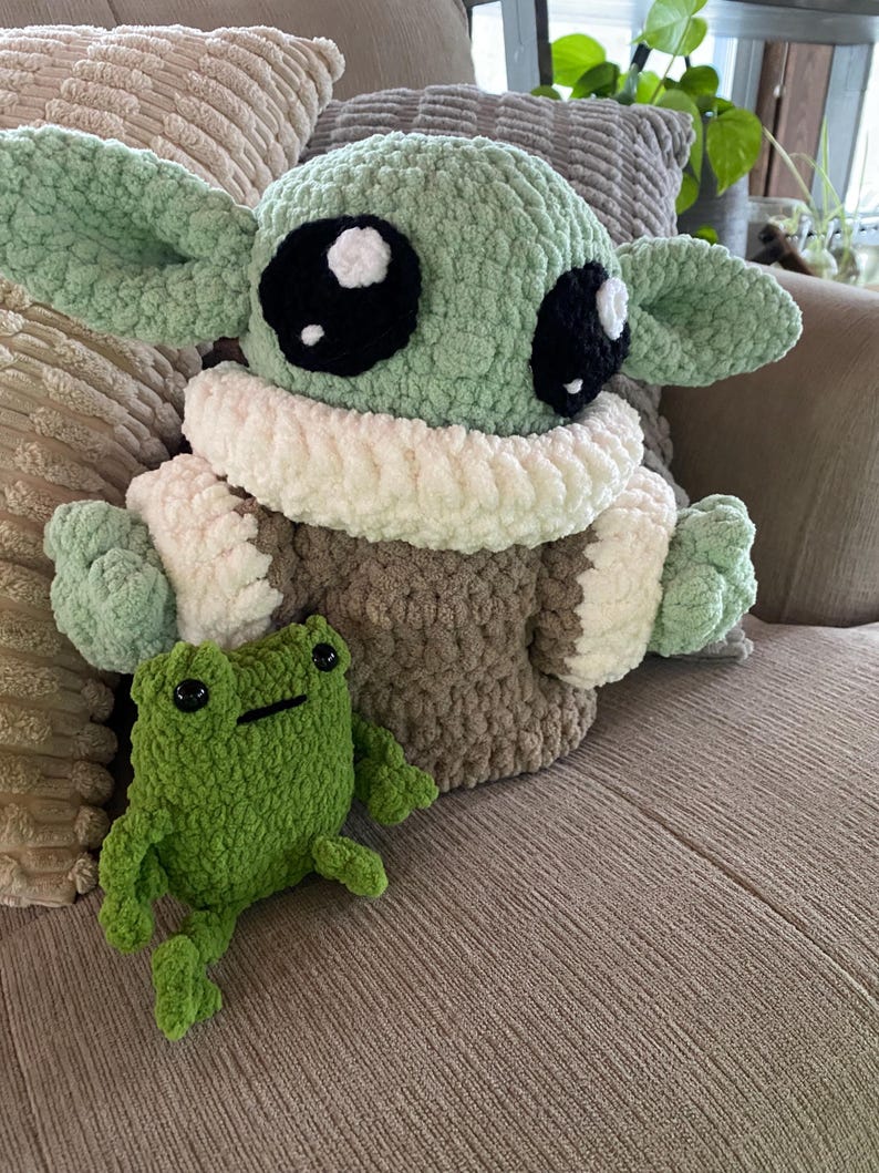 May include: A large, crocheted plush figure of a character with large black eyes, green skin, and large ears, wearing a brown and white outfit. A small green crocheted frog sits in front of the figure.