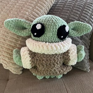 May include: A crocheted plush toy of a character with large black eyes, green skin, and large ears. The toy is wearing a brown and white outfit. The plush is resting on a couch with two decorative pillows.