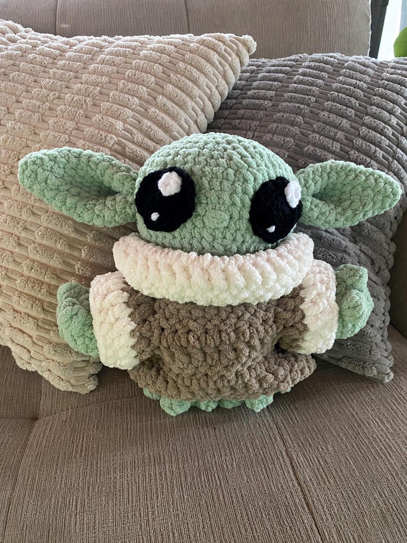 May include: A crocheted plush toy of a character with large black eyes, green skin, and large ears. The toy is wearing a brown and white outfit. The plush is resting on a couch with two decorative pillows.