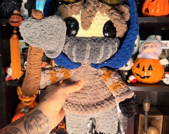 May include: A handmade crochet plush doll featuring a character with brown hair, wearing a blue hood, a gray face mask, and yellow-tinted goggles. The doll has black eyes and is holding a gray plush axe with a brown handle, wearing a gray sweater.