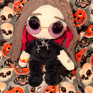 May include: A handmade crochet doll with a cream-colored face, dark brown hair, and round purple-tinted sunglasses. The doll wears a black shirt, black pants, and a layered silver-tone necklace with three cross pendants. It has white arms with flame-like orange and yellow patterns on one sleeve.