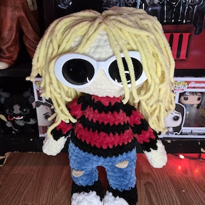 Könnte beinhalten: Handgefertigte Häkelpuppe mit gelbem Garnhaar, weißer Sonnenbrille und einem rot-schwarz gestreiften Pullover. Die Puppe trägt blaue Jeans mit zerrissenen Knien und schwarz-weiße Schuhe. Ein einzigartiger, handgefertigter Artikel.