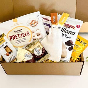 May include: A cardboard gift box filled with assorted snacks including a bag of yogurt covered pretzels, a Chuao honeycomb chocolate bar, Ferrero Rocher chocolates, a small white fabric pouch, Pepperidge Farm Milano cookies, and Stash tea.