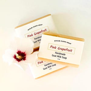 May include: Two rectangular bars of handmade goat milk soap labeled Pink Grapefruit by Sugar-Taffy-Soap, each weighing 4 oz. The soap bars are stacked and accompanied by a white hibiscus flower with a deep red center, all on a white background.