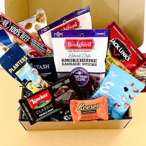 May include: A cardboard gift box filled with assorted snacks including Jack Link's beef jerky, Bridgford smokehouse sausage sticks, Planters salted cashews, Stash tea, Loacker double choc wafers, a Reese's peanut butter cup, and an energy nut mix.