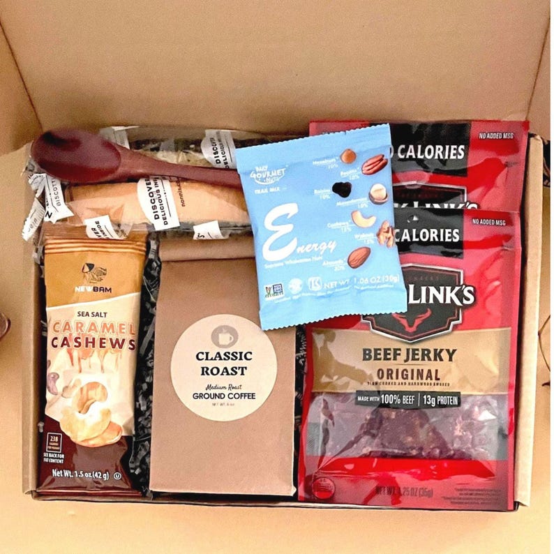 mens snack gift box filled with two beef jerky, classic roast ground coffee in a 4oz Kraft bag, trail mix, sea salt caramel cashews, wooden coffee spoon, and two wrapped iced biscotti.