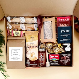 May include: A gift box containing two Nonni's biscotti, Coffee Delight candies, a bag of Cozy Classic Roast ground coffee, New Bam sea salt caramel cashews, a dark wooden spoon, a jar of Bee Harmony honey with a dipper, Stash Earl Grey tea, and Loacker Double Choc wafers.
