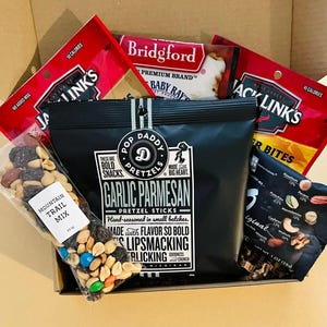 May include: A cardboard box filled with assorted snacks including a black bag of Pop Daddy Garlic Parmesan pretzel sticks, two red Jack Link's meat snack bags, a Bridgford Sweet Baby Ray's meat snack, a bag of nuts, and a clear bag of mountain trail mix.