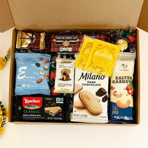 May include: A cardboard gift box filled with assorted snacks including a blue bag of trail mix, a package of Milano dark chocolate cookies, a bag of salted cashews, a Loacker dark chocolate wafer bar, shortbread, and two yellow Pukka tea bags.