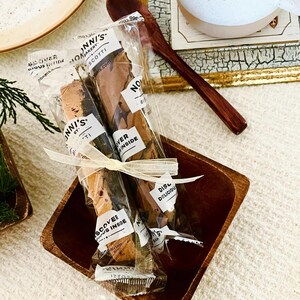 May include: Two Nonni's Bakery biscotti in clear plastic wrappers tied together with a thin cream-colored ribbon. The packaging features black text that reads Nonni's Bakery, Biscotti, and Discover Delicious Inside. The biscotti are brown.