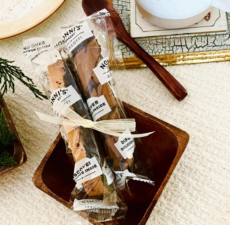 May include: Two Nonni's Bakery biscotti in clear plastic wrappers tied together with a thin cream-colored ribbon. The packaging features black text that reads Nonni's Bakery, Biscotti, and Discover Delicious Inside. The biscotti are brown.