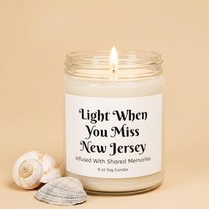 May include: A clear glass jar containing a white soy candle with a lit wick. The white label features black text reading Light When You Miss New Jersey, Infused With Shared Memories, 9 oz Soy Candle. Two seashells are placed in front of the jar.