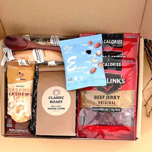 mens snack gift box filled with two beef jerky, classic roast ground coffee in a Kraft bag, trail mix, sea salt caramel cashews, wooden coffee spoon, and two wrapped iced biscotti.