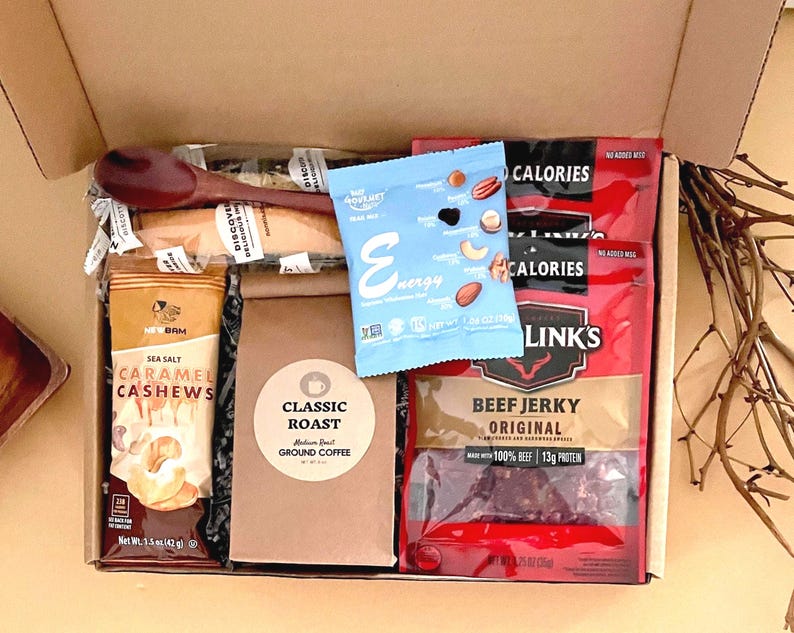 mens snack gift box filled with two beef jerky, classic roast ground coffee in a Kraft bag, trail mix, sea salt caramel cashews, wooden coffee spoon, and two wrapped iced biscotti.