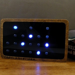 Op de afbeelding: Een houten frame met een zwarte achtergrond en een raster van blauwe lampjes. De lampjes zijn in een patroon gerangschikt en het frame heeft de tekst "Peculiar Devices" onderaan.