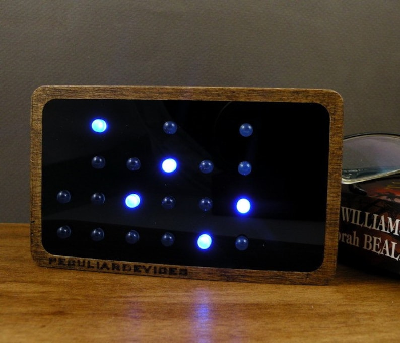 Op de afbeelding: Een houten frame met een zwarte achtergrond en een raster van blauwe lampjes. De lampjes zijn in een patroon gerangschikt en het frame heeft de tekst "Peculiar Devices" onderaan.
