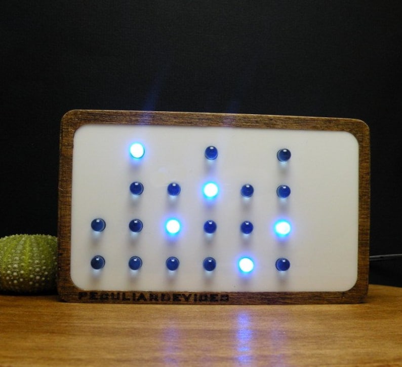 Op de afbeelding: Een rechthoekige witte klok met een houten frame. De wijzerplaat van de klok heeft een raster van blauwe en witte lampjes. De wijzerplaat van de klok heeft ook de tekst "PECULIARDDEVICES" onderaan.