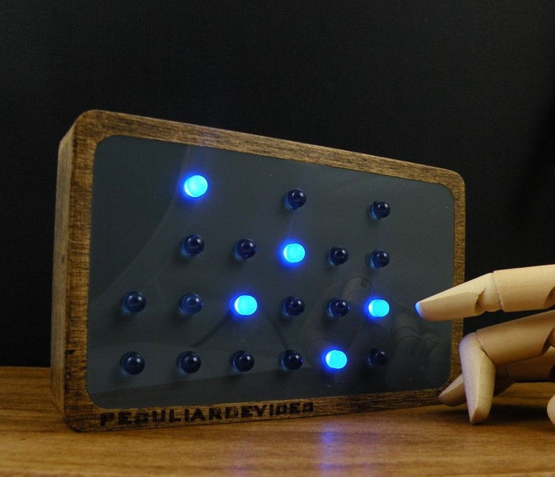 Op de afbeelding: Een houten frame met een donkergrijze achtergrond en een raster van blauwe lampjes. De lampjes zijn in een driehoekige vorm gerangschikt. Het frame heeft de tekst "PECULIARDDEVICES" gegraveerd aan de onderkant.