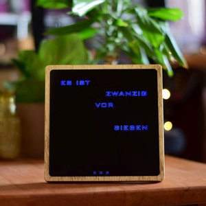 May include: A wooden clock with a black face and blue digital display showing the time "ES IST ZWANZIG VOR SIEBEN".