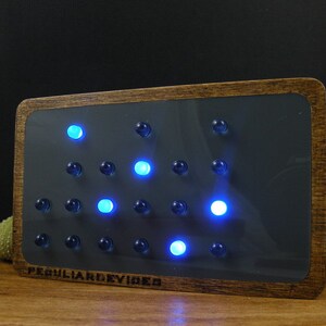 Op de afbeelding: Een houten ingelijste klok met een donkergrijze achtergrond en een raster van blauwe en witte LED-lampjes. De klok geeft de tijd 12:00 aan. De klok is gelabeld met "Peculiar Devices".