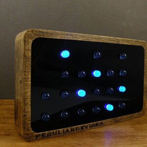 Op de afbeelding: Een houten ingelijste klok met een zwarte achtergrond en blauwe LED-verlichting. De klok geeft de tijd weer met behulp van een stippenmatrixpatroon. De klok is gelabeld met "Peculiar Devices".