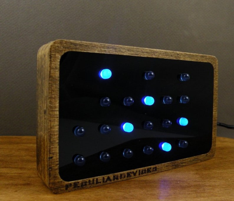 Op de afbeelding: Een houten ingelijste klok met een zwarte achtergrond en blauwe LED-verlichting. De klok geeft de tijd weer met behulp van een stippenmatrixpatroon. De klok is gelabeld met "Peculiar Devices".