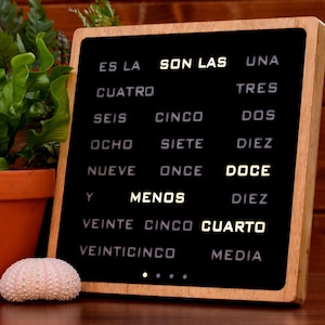 Puede incluir: Un letrero de madera con marco negro y texto blanco sobre fondo negro. El texto dice "ES LA SON LAS UNA CUATRO TRES SEIS CINCO DOS OCHO SIETE DIEZ NUEVE ONCE DOCE Y MENOS DIEZ VEINTE CINCO CUARTO VEINTICINCO MEDIA".