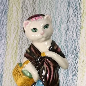 Könnte beinhalten: Keramikfigur einer Katze mit Schal und blauem Rock, die einen gelben Korb hält. Die Katze hat grüne Augen und eine rosa Schleife im Haar. Die Figur steht auf einem steinartigen Sockel.