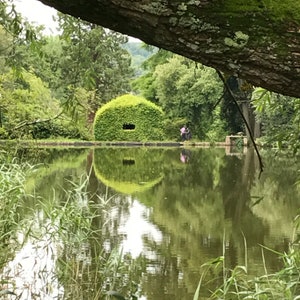 May include: A serene outdoor scene featuring a pond reflecting lush greenery and a unique, rounded structure covered in dense foliage. A tree branch frames the top, and a couple stands near the water's edge.