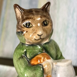 May include: A ceramic figurine of a cat with brown tabby markings on its head, wearing a green jacket over an orange garment. The cat is holding a small, light grey, rounded pot with both hands. The surface of the figurine is glossy and smooth.
