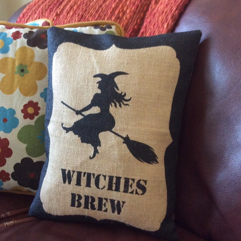 May include: A rectangular burlap pillow with a black witch silhouette riding a broom. The pillow has a black border and the words "WITCHES BREW" in black block letters. The pillow is a Halloween-themed decorative item.