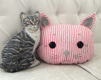 May include: Two plush cat pillows, one gray and white tabby cat and one pink cat with white stripes. The pink cat has black eyes and a gray nose.