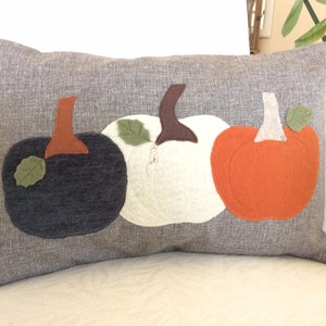 May include: A rectangular grey cushion featuring three felt pumpkins. The pumpkins are black, white, and orange, each with a brown stem and green leaf. The cushion is on a white surface.