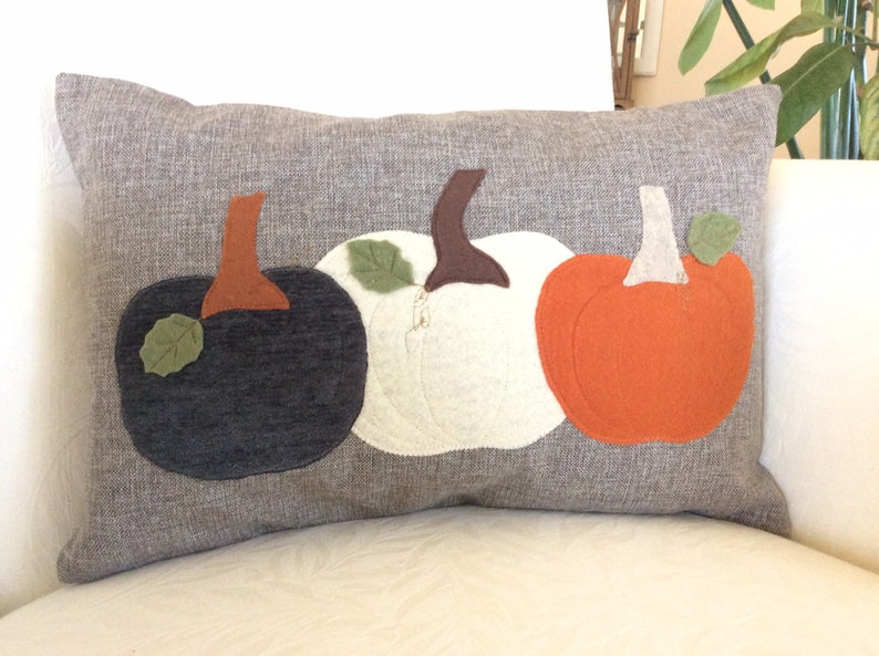 May include: A rectangular grey cushion featuring three felt pumpkins. The pumpkins are black, white, and orange, each with a brown stem and green leaf. The cushion is on a white surface.