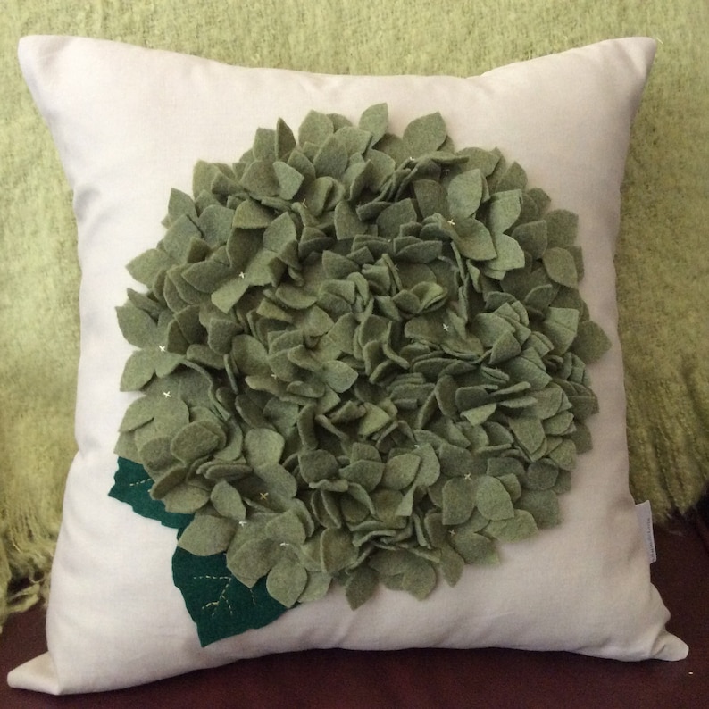 May include: A white pillow with a large green felt flower appliqu&eacute;. The flower is made up of many individual petals and has a green felt leaf at the bottom.