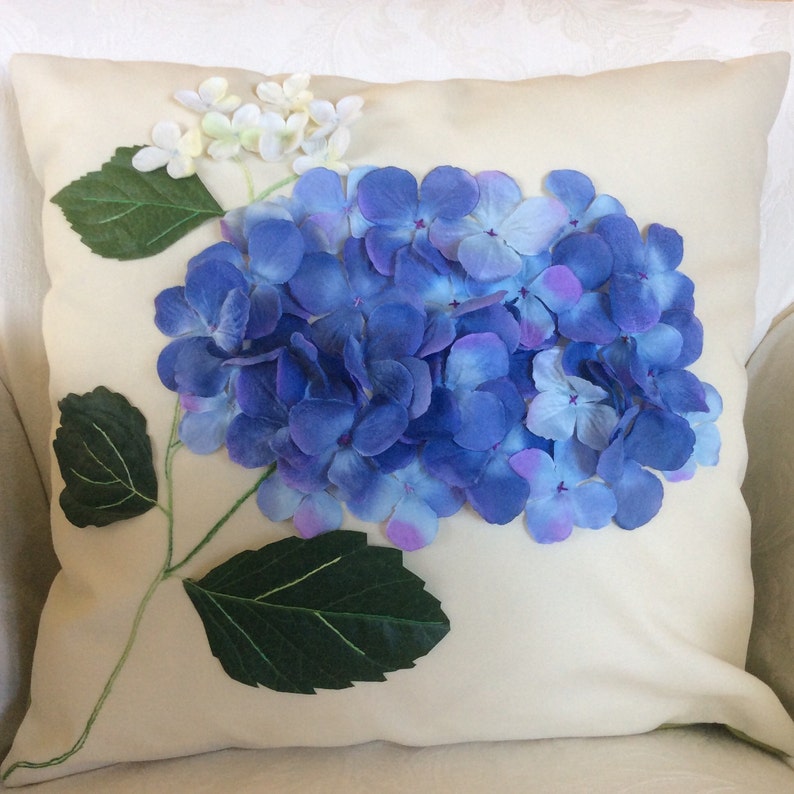 May include: A white pillow with a blue and purple hydrangea flower appliqu&eacute;. The flower is made of fabric and has green leaves.