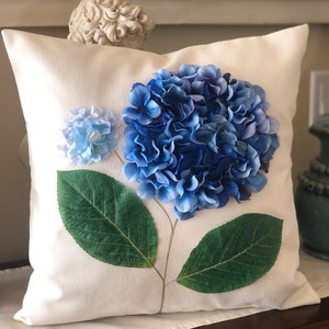 May include: A white pillow with a blue and green hydrangea flower design. The flower is made of fabric and has a realistic appearance.
