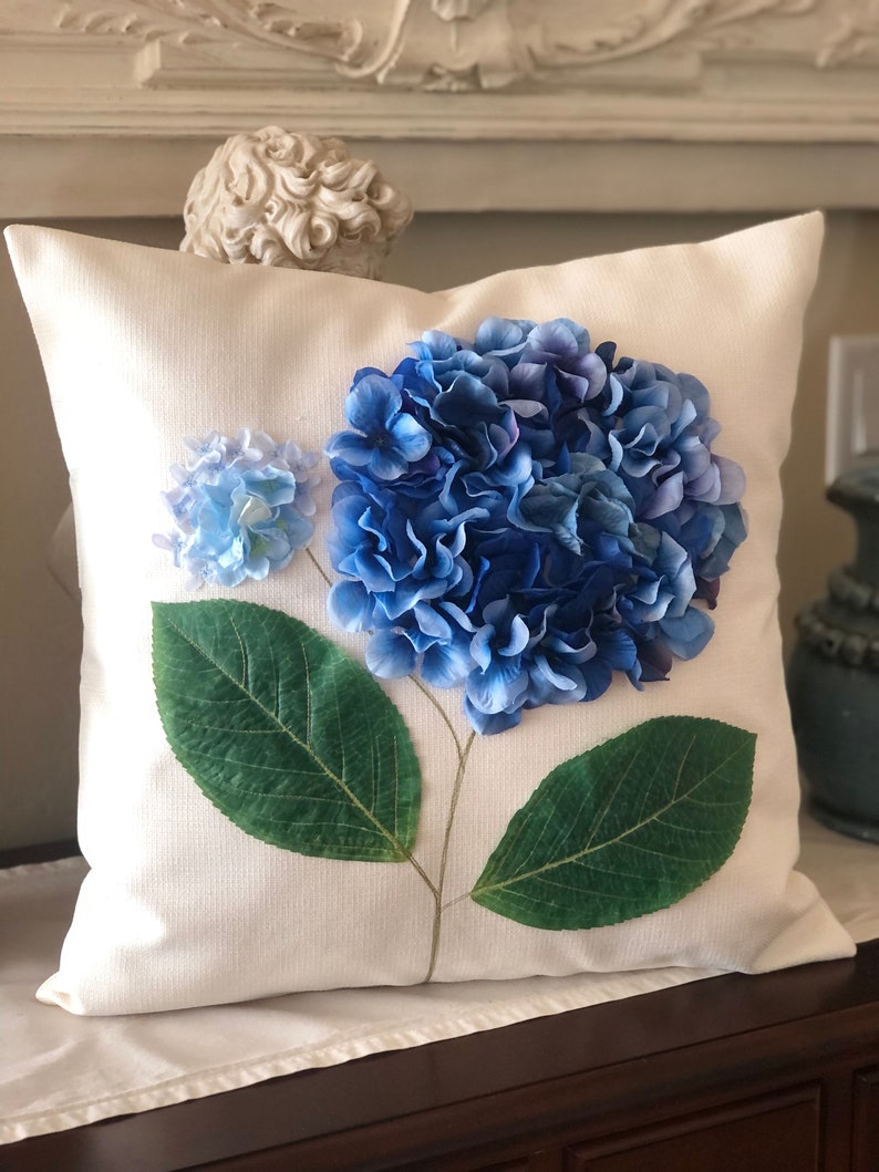 May include: A white pillow with a blue and green hydrangea flower design. The flower is made of fabric and has a realistic appearance.
