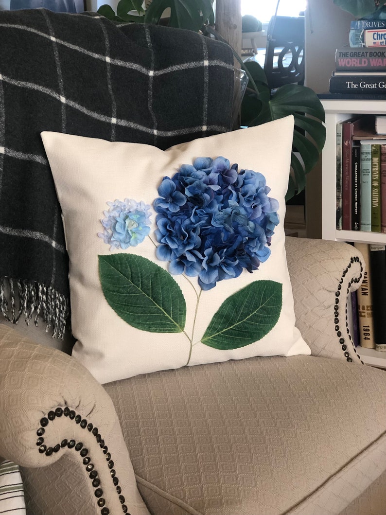 May include: A white pillow with a blue and green hydrangea flower pressed onto the fabric.