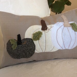 May include: A rectangular cushion featuring three appliqued pumpkins in white and dark grey, with brown stalks and green leaves, against a tan linen-effect background. The cushion is on a white patterned surface.