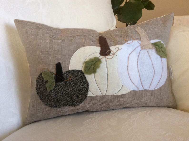 May include: A rectangular cushion featuring three appliqued pumpkins in white and dark grey, with brown stalks and green leaves, against a tan linen-effect background. The cushion is on a white patterned surface.