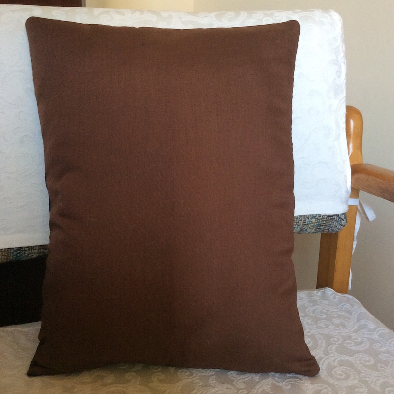 May include: A solid brown square pillow. The pillow is made of a textured fabric and is placed on a chair with a white patterned cushion cover. The pillow is a warm, earthy tone, suitable for home decor.