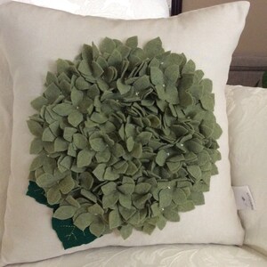 May include: A white pillow with a green felt hydrangea flower design. The flower is made of individual felt petals.