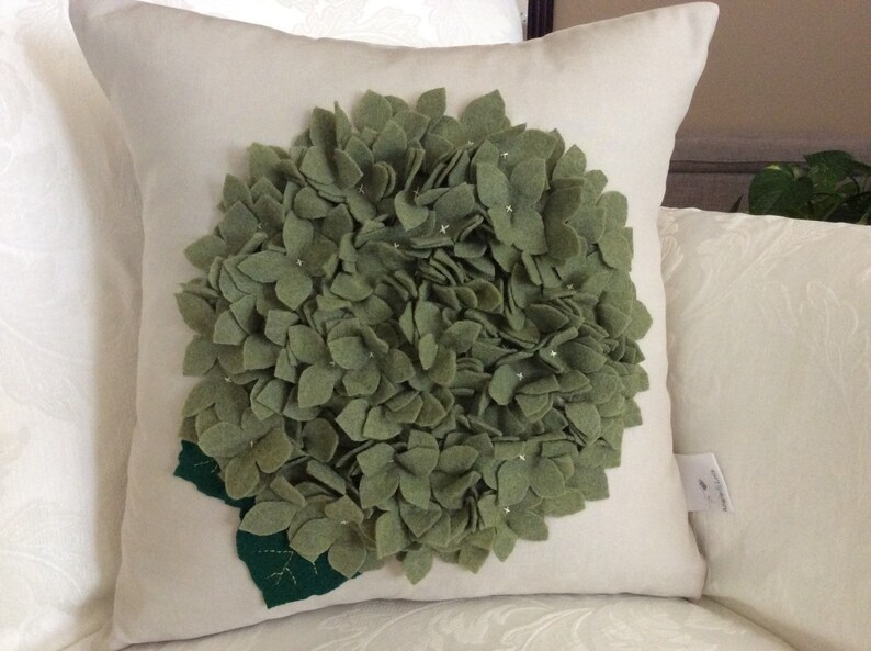 May include: A white pillow with a green felt hydrangea flower design. The flower is made of individual felt petals.