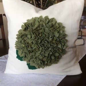 May include: A white pillow with a green felt flower appliqu&eacute;. The flower is made of many small, ruffled petals and has a green felt stem.