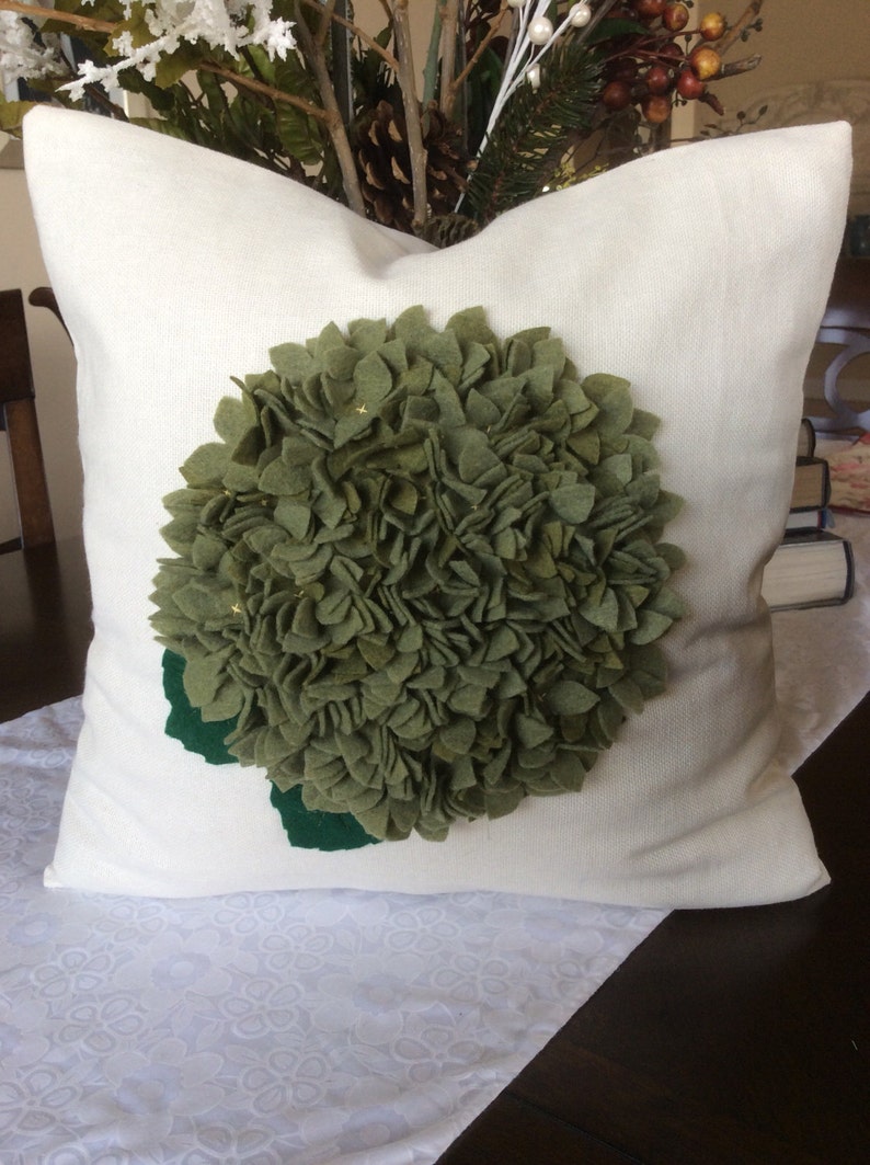 May include: A white pillow with a green felt flower appliqu&eacute;. The flower is made of many small, ruffled petals and has a green felt stem.